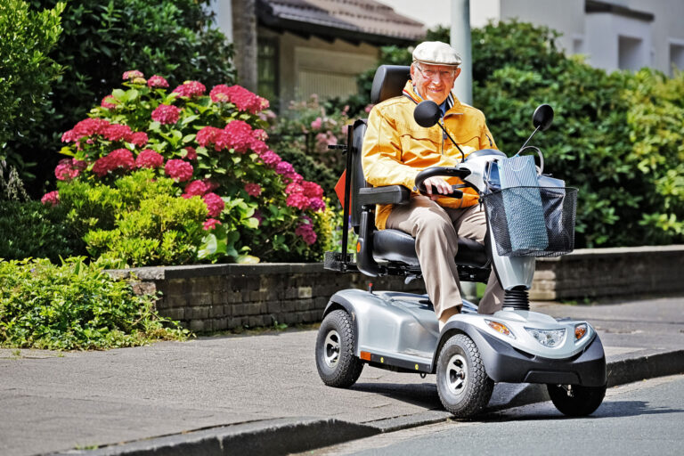 Älterer Mann mit Mütze und gelber Jacke fährt fröhlich auf einem silbernen Elektromobil auf der Straße, im Hintergrund blühende Hortensien und Grünpflanzen.