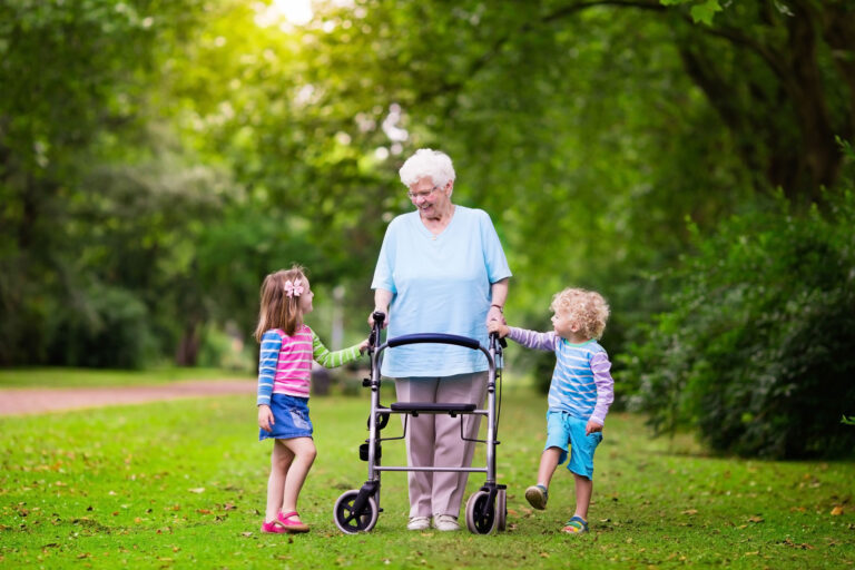 Ältere Frau mit Rollator geht im Park, begleitet von zwei kleinen Kindern, die jeweils eine ihrer Hände halten. Alle wirken fröhlich und entspannt.