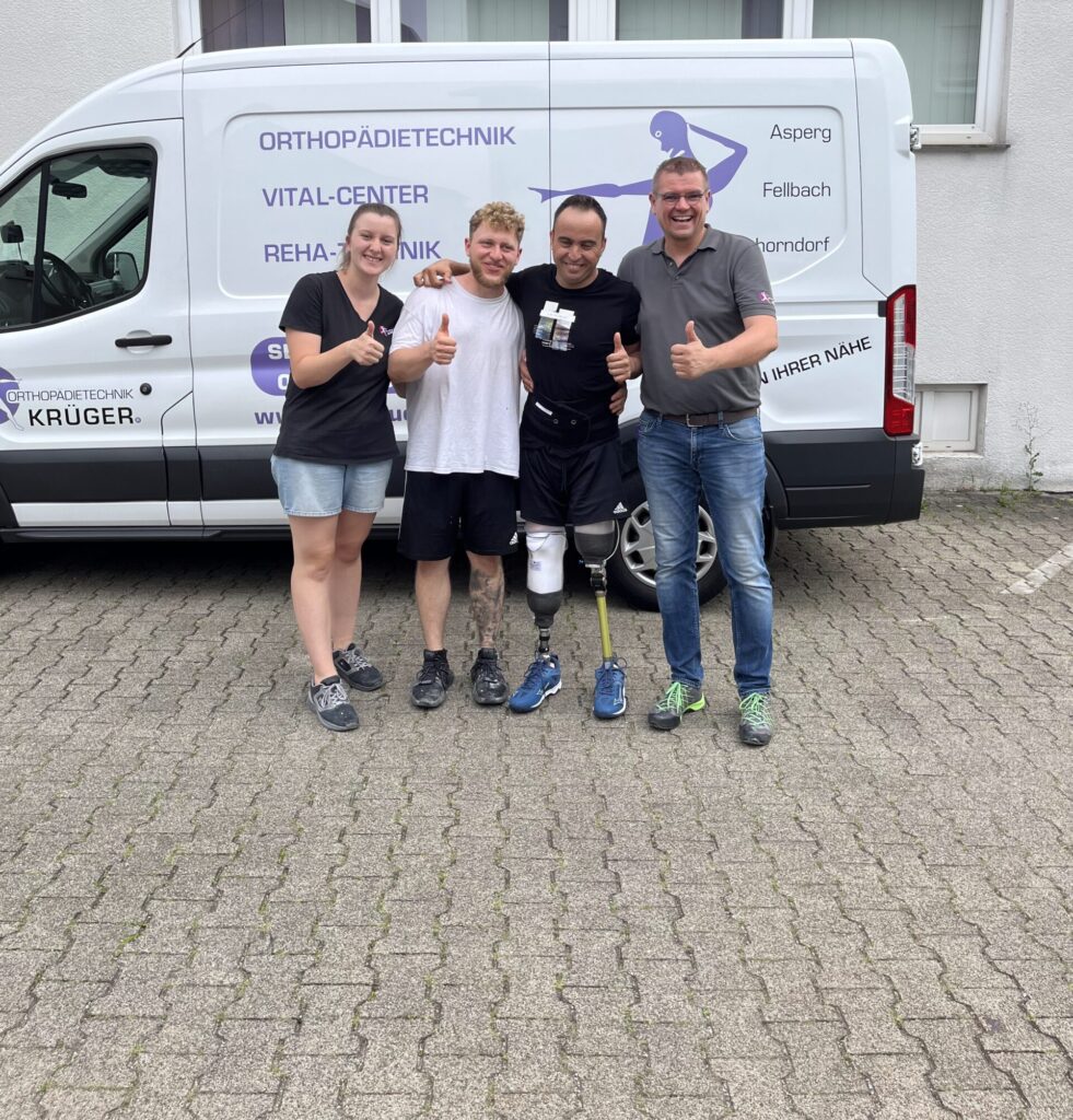 Vier Personen stehen lächelnd vor einem Transporter mit der Aufschrift 'Orthopädietechnik Krüger'. In der Mitte ein Mann mit Beinprothese, alle vier zeigen Daumen hoch und wirken gut gelaunt