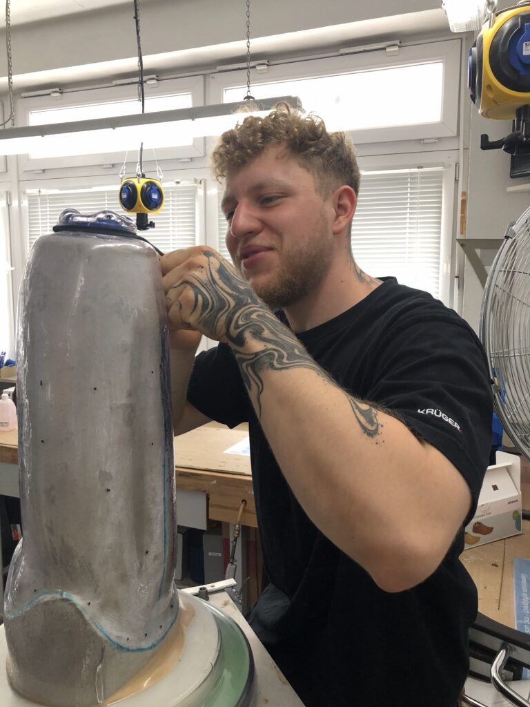 Ein Orthopädietechniker arbeitet konzentriert an einer Prothesenform in der Werkstatt. Er trägt ein schwarzes T-Shirt und hat tätowierte Arme.