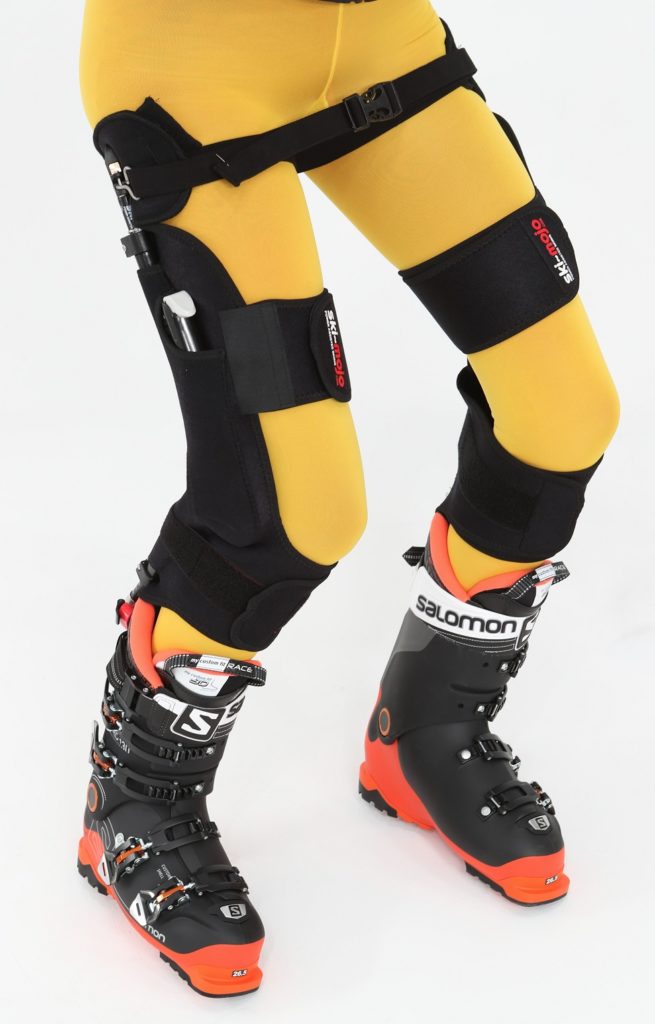 Beine mit gelber Skibekleidung, darauf schwarze orthopädische Knieorthesen der Marke Ortema, getragen zusammen mit schwarzen Skischuhen von Salomon.